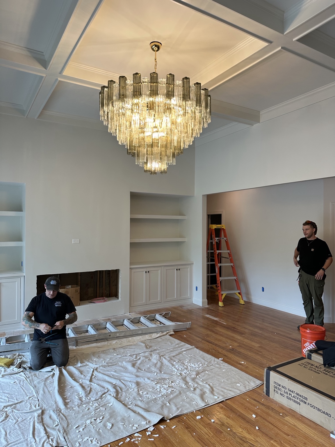 Scott, electrician, and Ash, project manager, beneath a large glass chandelier during installation in a room with a custom coffered ceiling