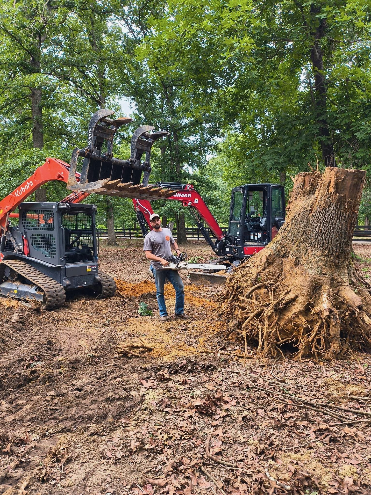Chase standing with a chainsaw beside a large removed stump and compact excavation equipment